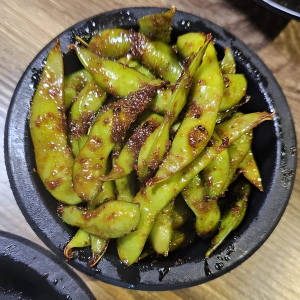 A black bowl filled with bright green, glazed edamame pods, ready to be served at Slurpin' Ramen Bar.