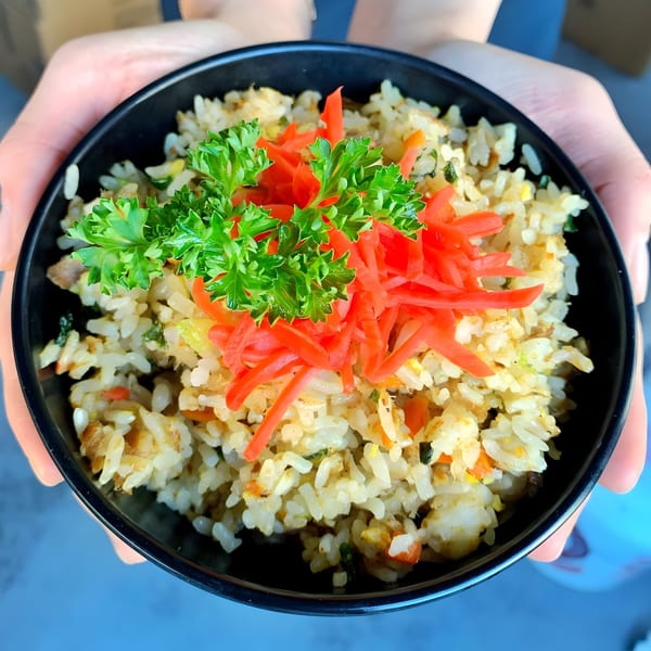 Hands holding a black bowl of fried rice topped with red pickled ginger and green parsley.