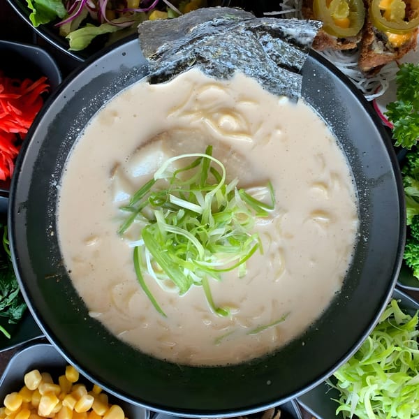 Creamy ramen with green onions and nori in a black bowl, surrounded by various toppings.