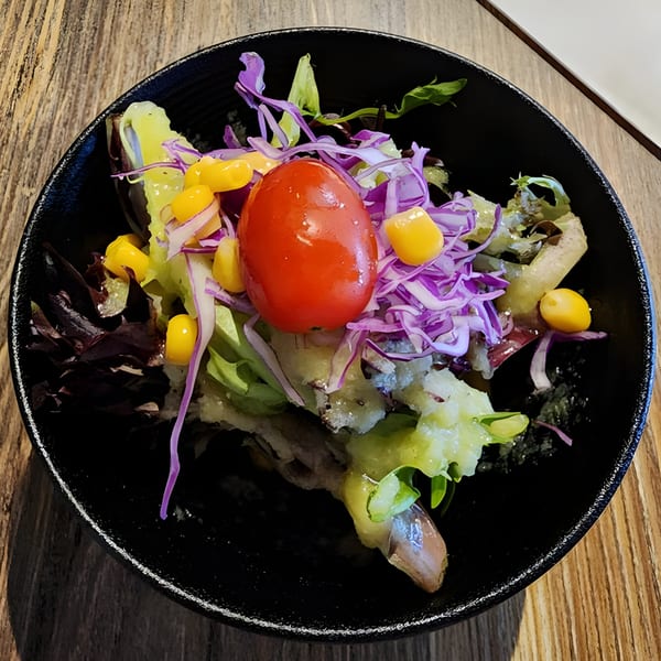 A vibrant side salad with lettuce, purple cabbage, corn, and a cherry tomato in a black bowl.